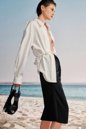 Woman on a beach wearing a white shirt and black skirt, holding black high heel shoes, with a blue ocean and sky background.