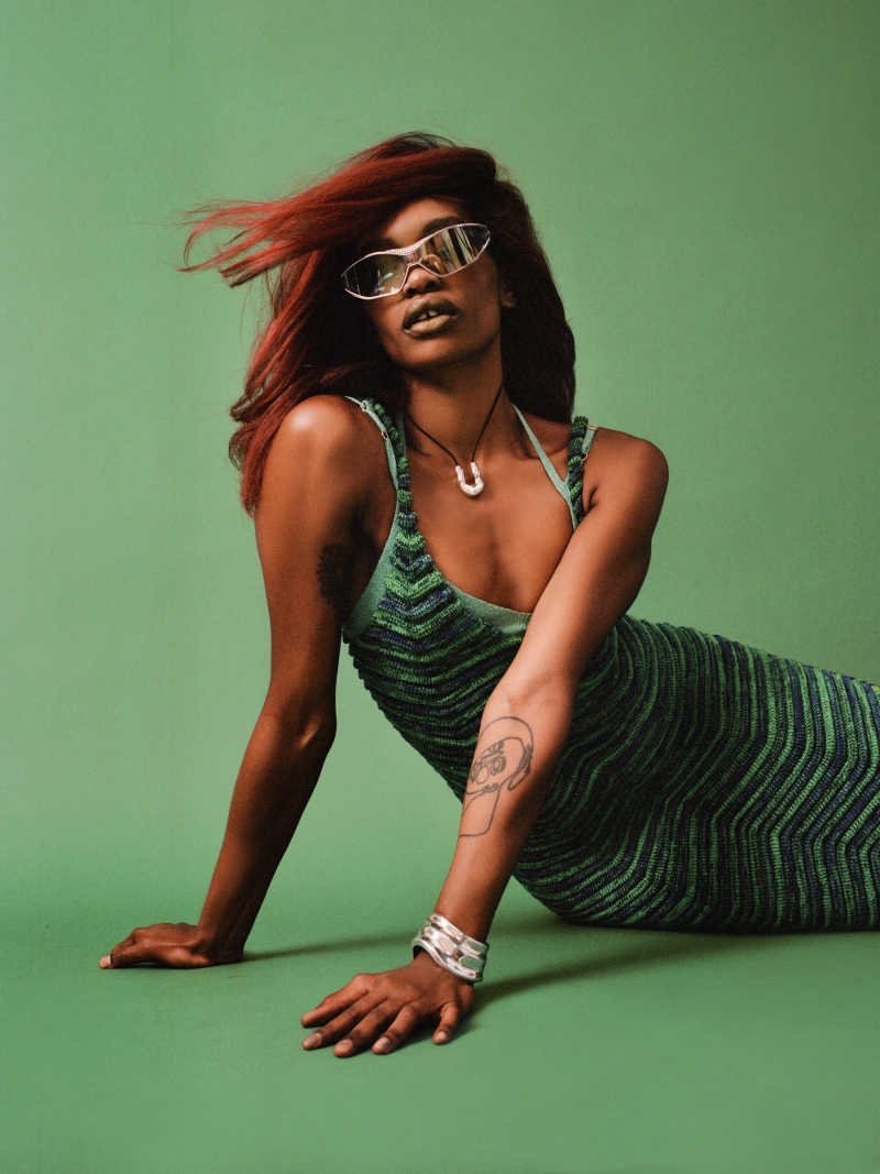 Portrait of a Black woman with red hair wearing sunglasses and a knitted dress, posing on a green background in a studio setting.