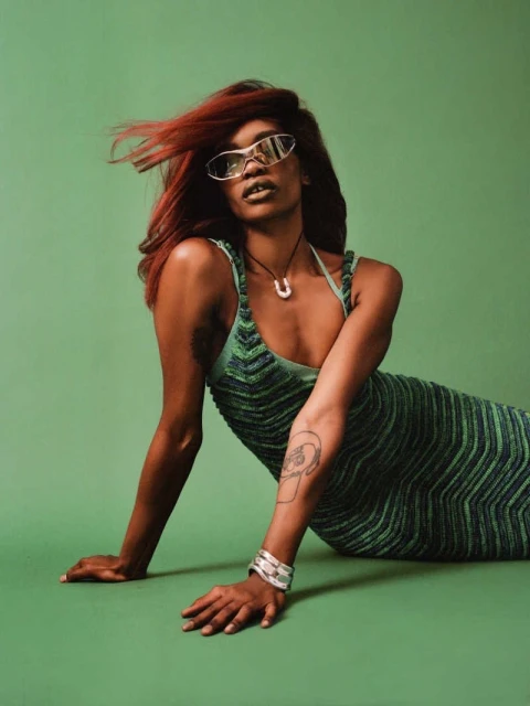 Portrait of a Black woman with red hair wearing sunglasses and a knitted dress, posing on a green background in a studio setting.