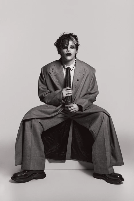 Person in an oversized grey suit and tie seated against a plain neutral backdrop in a studio fashion portrait.