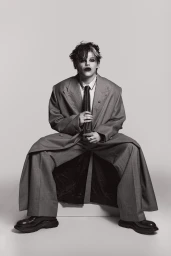 Person in an oversized grey suit and tie seated against a plain neutral backdrop in a studio fashion portrait.