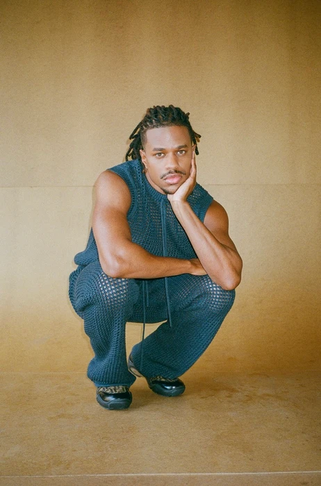 Man with dreadlocks wearing dark blue knit sleeveless top and matching pants, squatting against a plain tan wall.