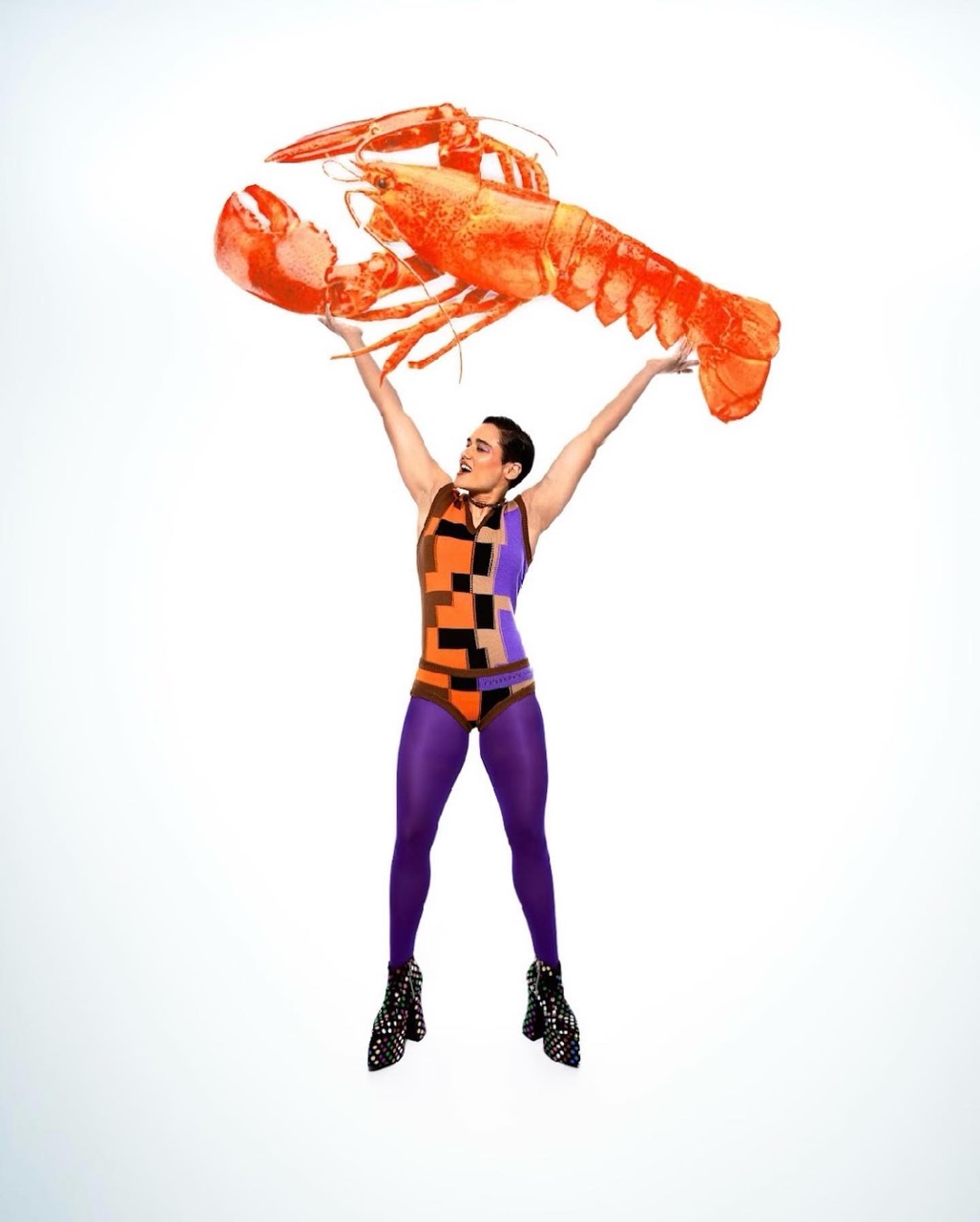 Woman in patterned bodysuit and tights holding a large orange lobster overhead against a white background in a studio setting.