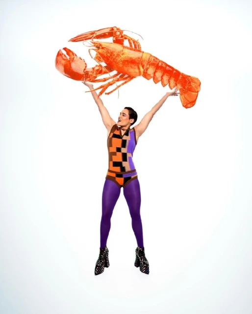 Woman in patterned bodysuit and tights holding a large orange lobster overhead against a white background in a studio setting.