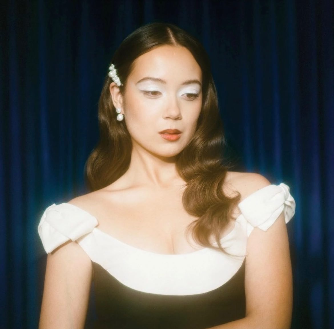 Portrait of a woman with dramatic white eyeshadow, red lipstick, and a black and white dress against a dark blue background.