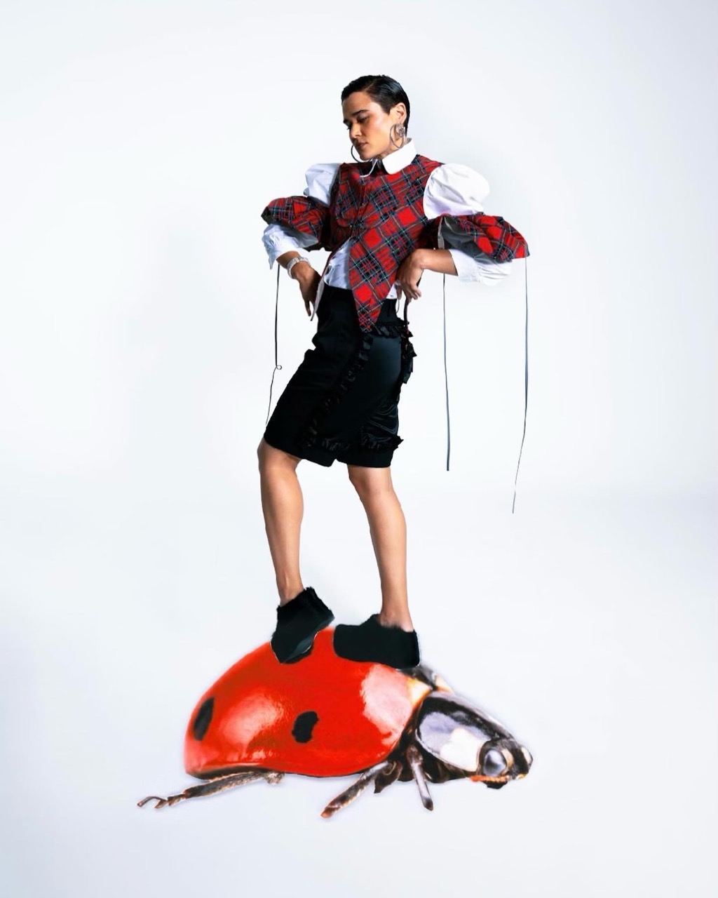 Fashion photo of a woman in plaid top and black skirt standing on a ladybug against a white background in a studio setting.