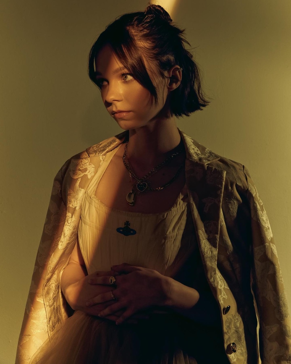 Portrait of a young woman with short brown hair wearing a cream-colored dress and gold blazer, posing indoors with neutral background.