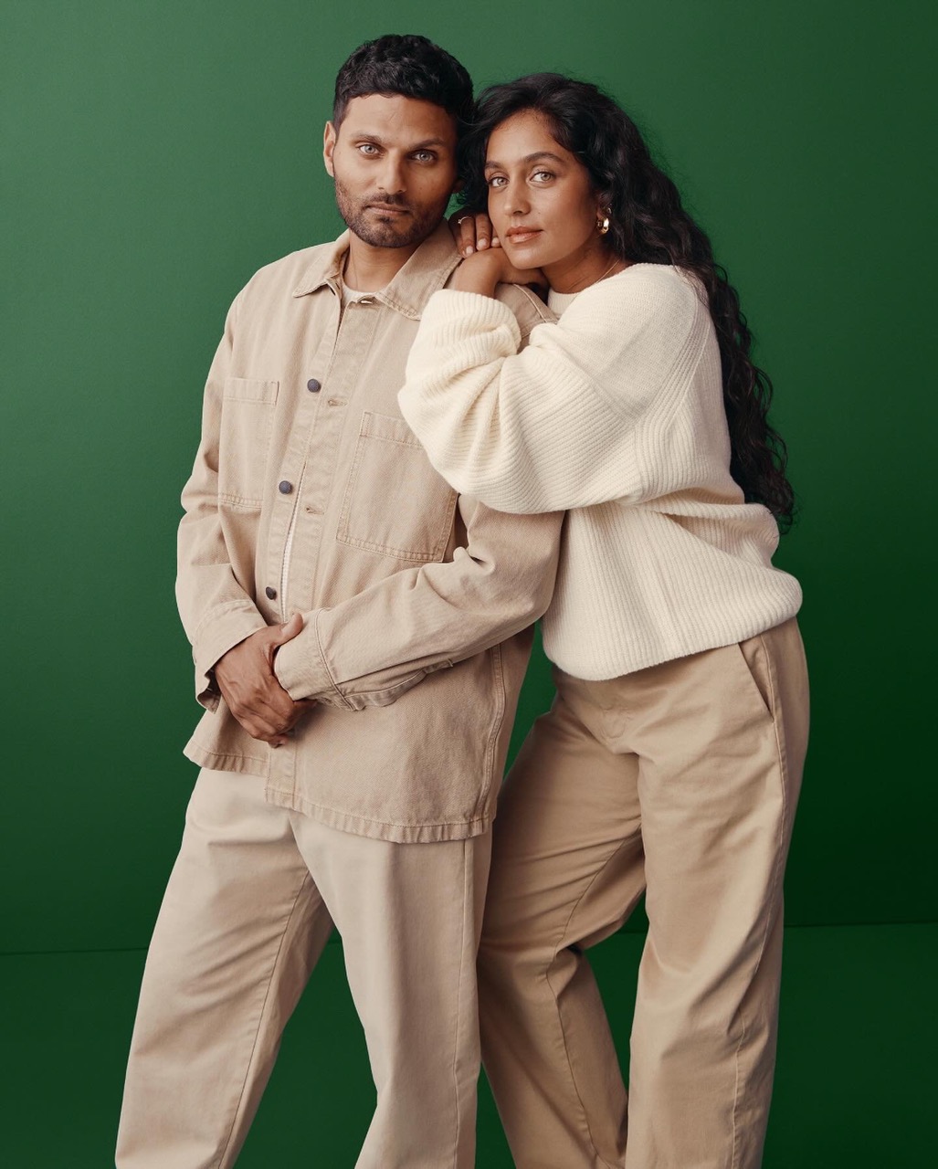 Portrait of a couple in matching beige outfits against a solid green background. The woman rests her head on the man's shoulder.