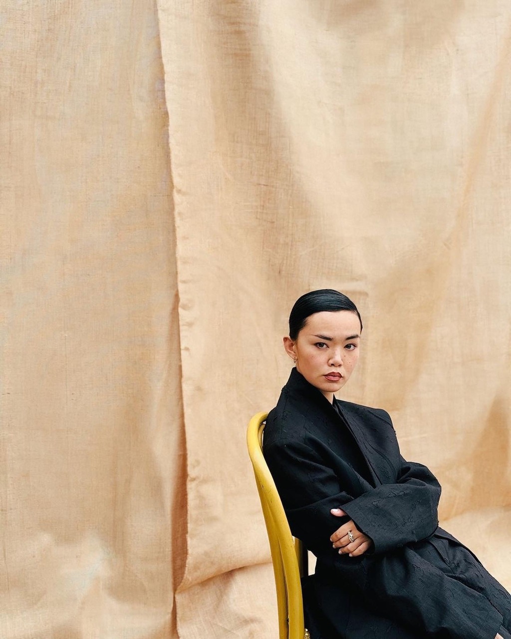 Portrait of an adult Asian woman wearing a black wrap blazer sitting on a yellow chair against a beige fabric backdrop.