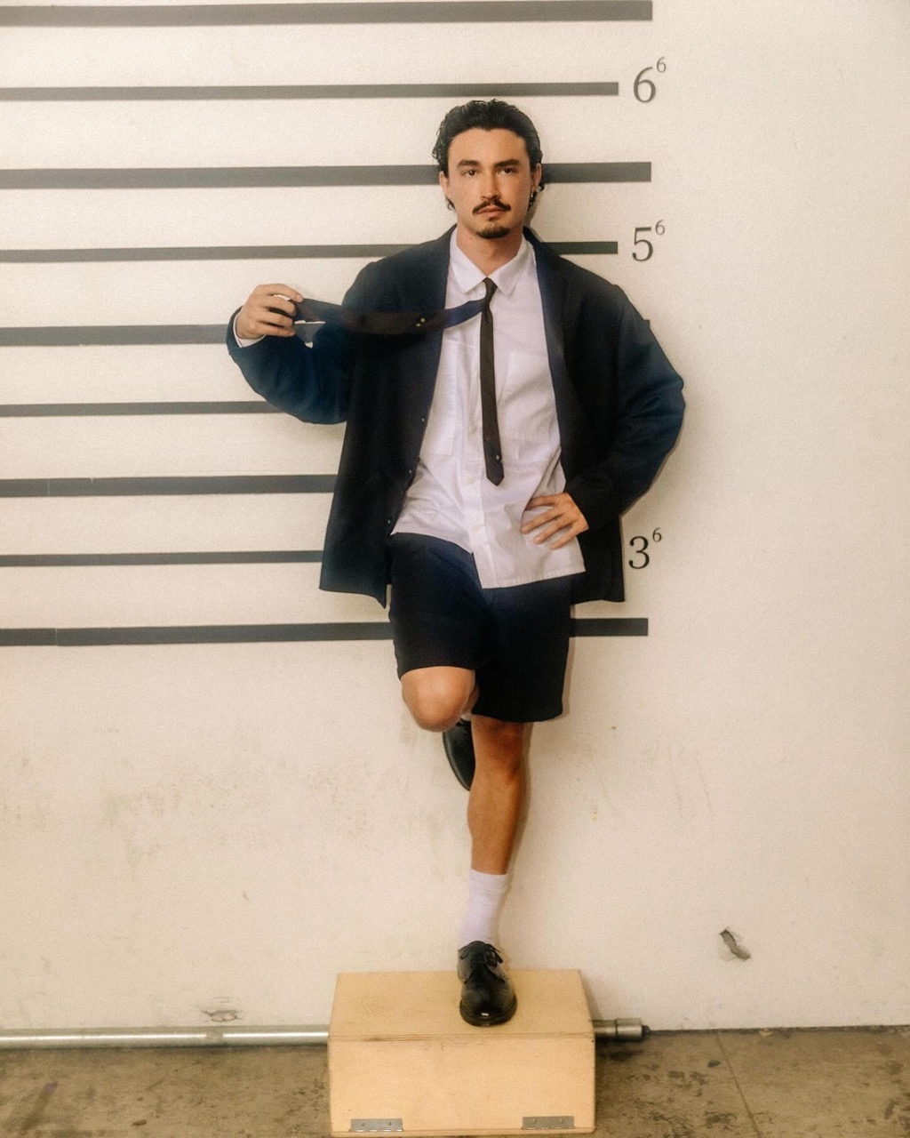 Man in a suit and shorts stands on a box in front of a height chart, holding a tie. He has dark hair and a mustache.