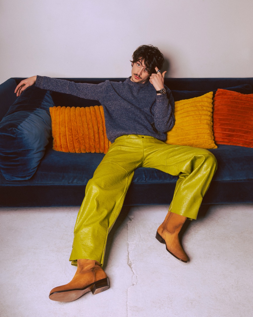 Young man in a gray sweater and bright green leather pants sits on a blue velvet couch with orange and blue throw pillows.