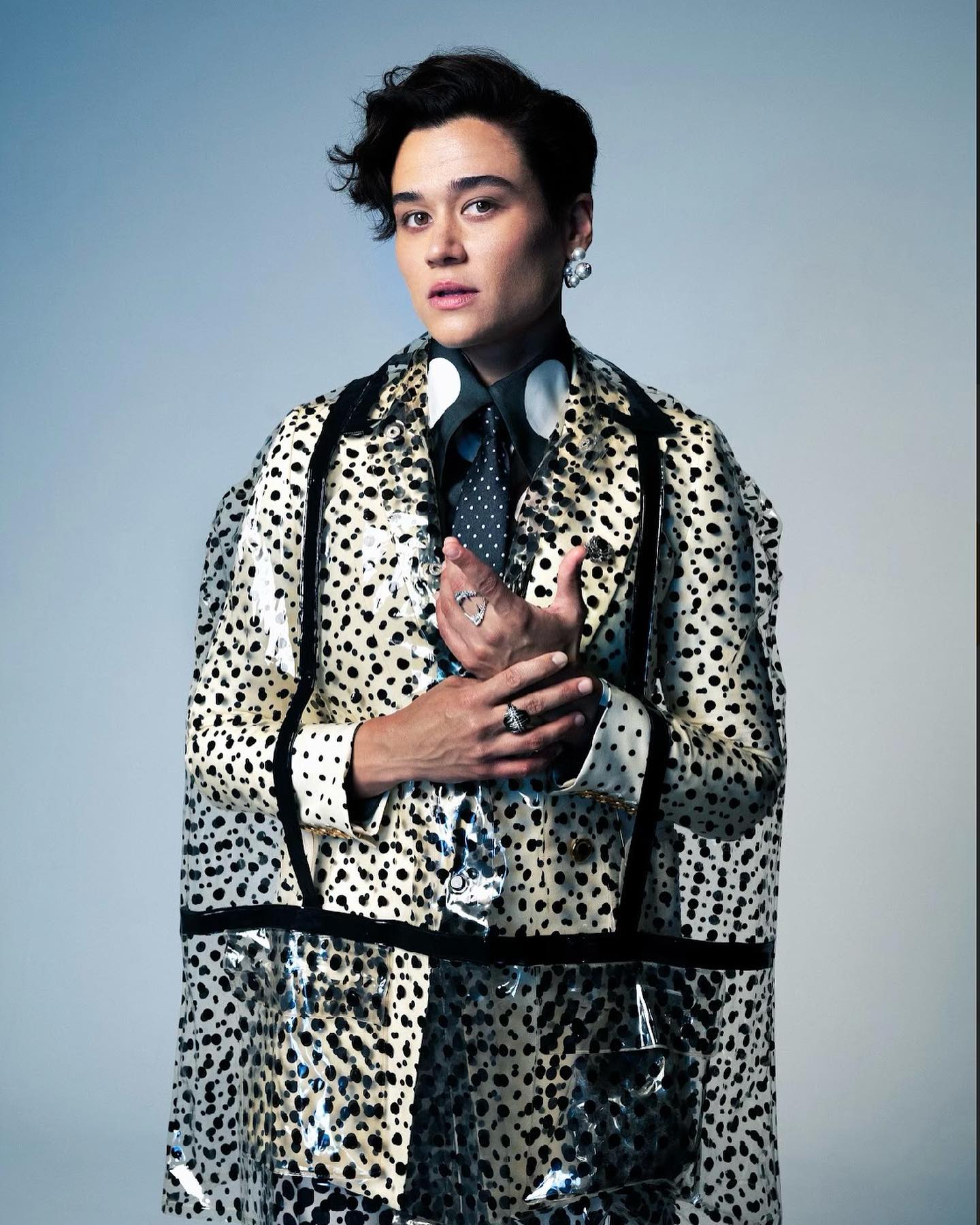 Person with dark hair poses wearing a transparent, spotted raincoat, dark shirt, and tie against a light blue-grey backdrop.