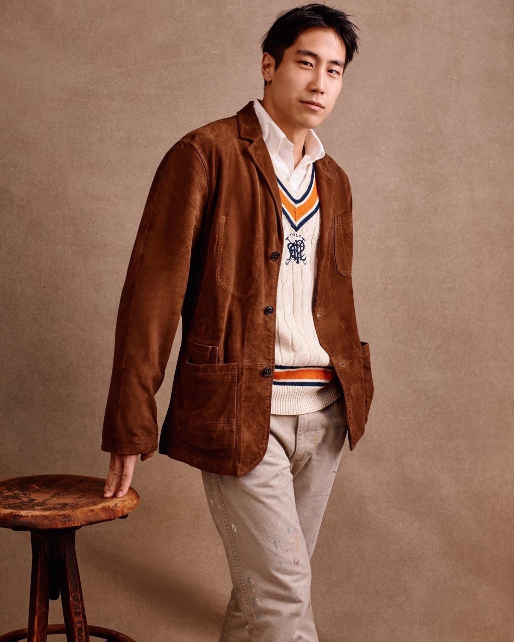 Adult man in white collared shirt, knit sweater, and brown suede jacket poses near rustic stool against neutral backdrop.
