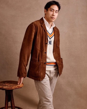 Adult man in white collared shirt, knit sweater, and brown suede jacket poses near rustic stool against neutral backdrop.