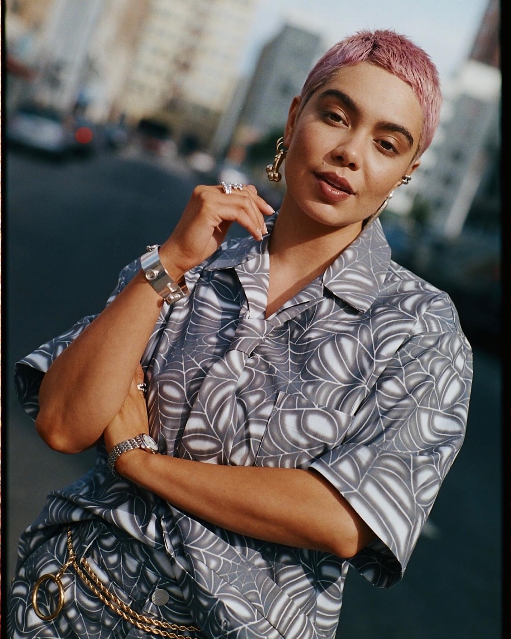 Stylish woman in patterned shirt on city street, accessorized with jewelry. Buildings and a car are blurred in the background.