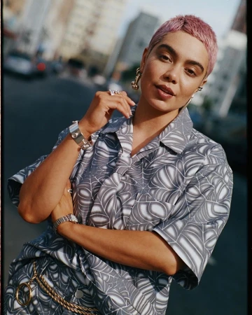 Stylish woman in patterned shirt on city street, accessorized with jewelry. Buildings and a car are blurred in the background.
