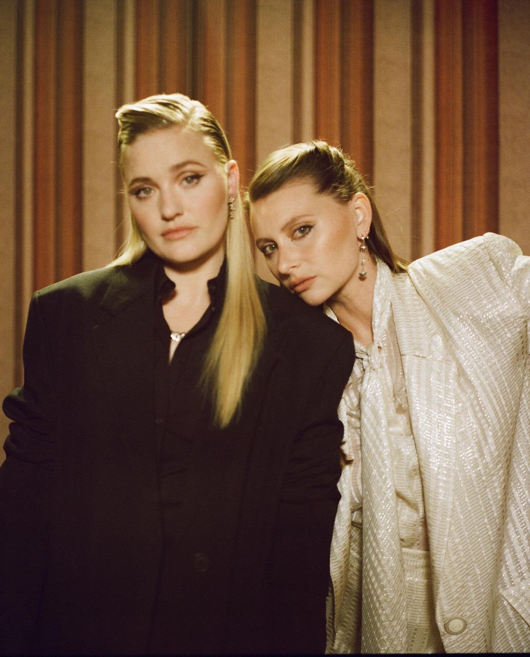 Two women pose indoors against a striped wall. One woman wears a black blazer, the other a silver sequined outfit, both with sleek hair.