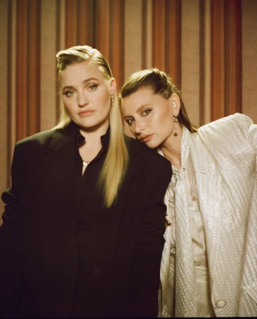 Two women pose indoors against a striped wall. One woman wears a black blazer, the other a silver sequined outfit, both with sleek hair.