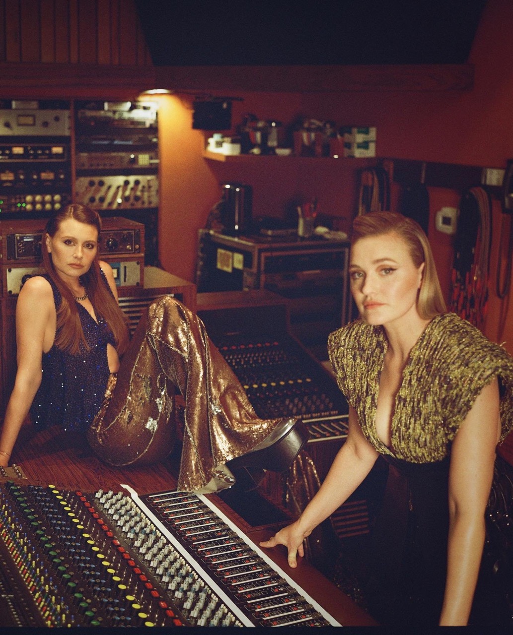 Two women in a music studio with vintage equipment. One is sitting by the soundboard wearing gold pants and a sparkly top.