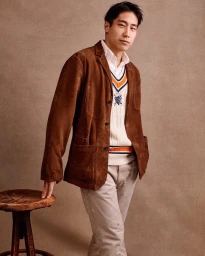 Man wearing a brown suede jacket over a cream V-neck sweater and light grey trousers standing in a studio