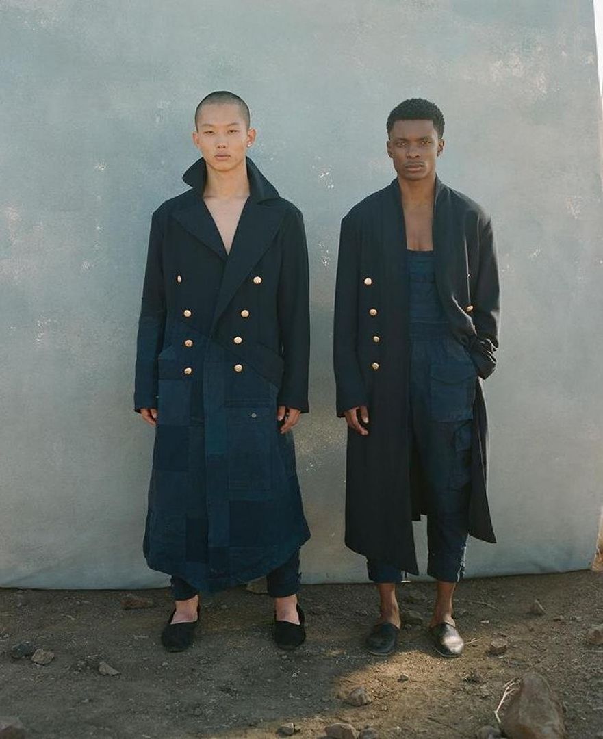 Two fashion models stand side by side outdoors in a neutral setting, wearing stylish navy blue coats and coordinating outfits.