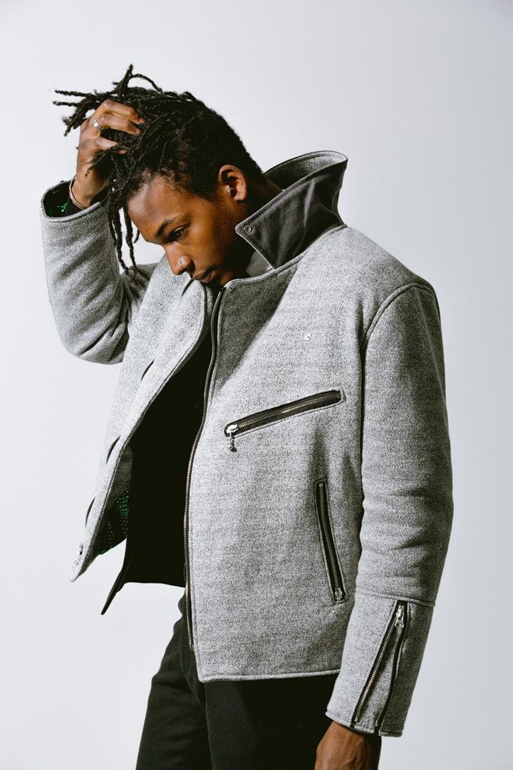 Studio shot of a Black male model in a grey jacket with black accents, hand to head, against a gradient white background.
