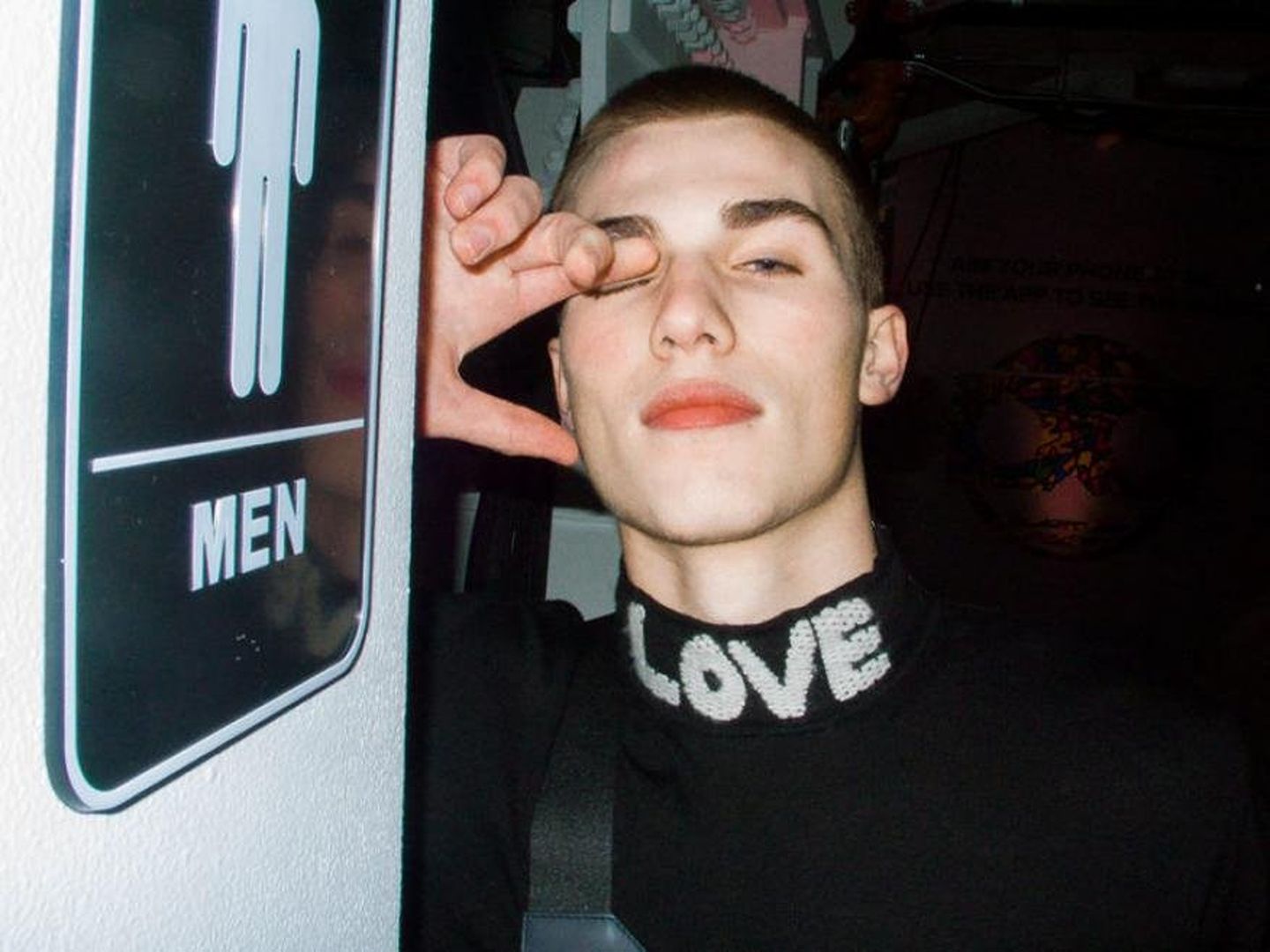 Close-up shot of a young adult male with a buzz cut wearing a black turtleneck sweater with the word LOVE on the collar indoors.