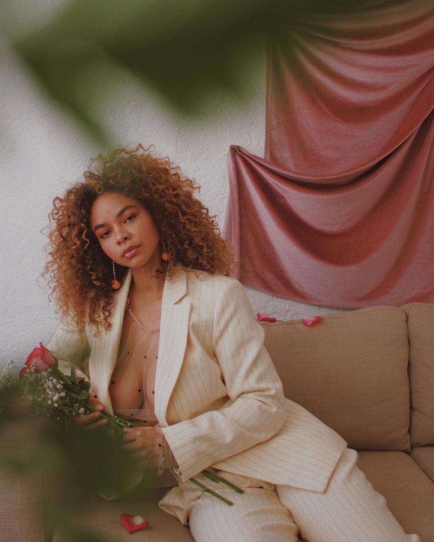 A young woman with curly hair poses indoors wearing a cream suit, holding pink roses on a couch with a blush pink draped background.