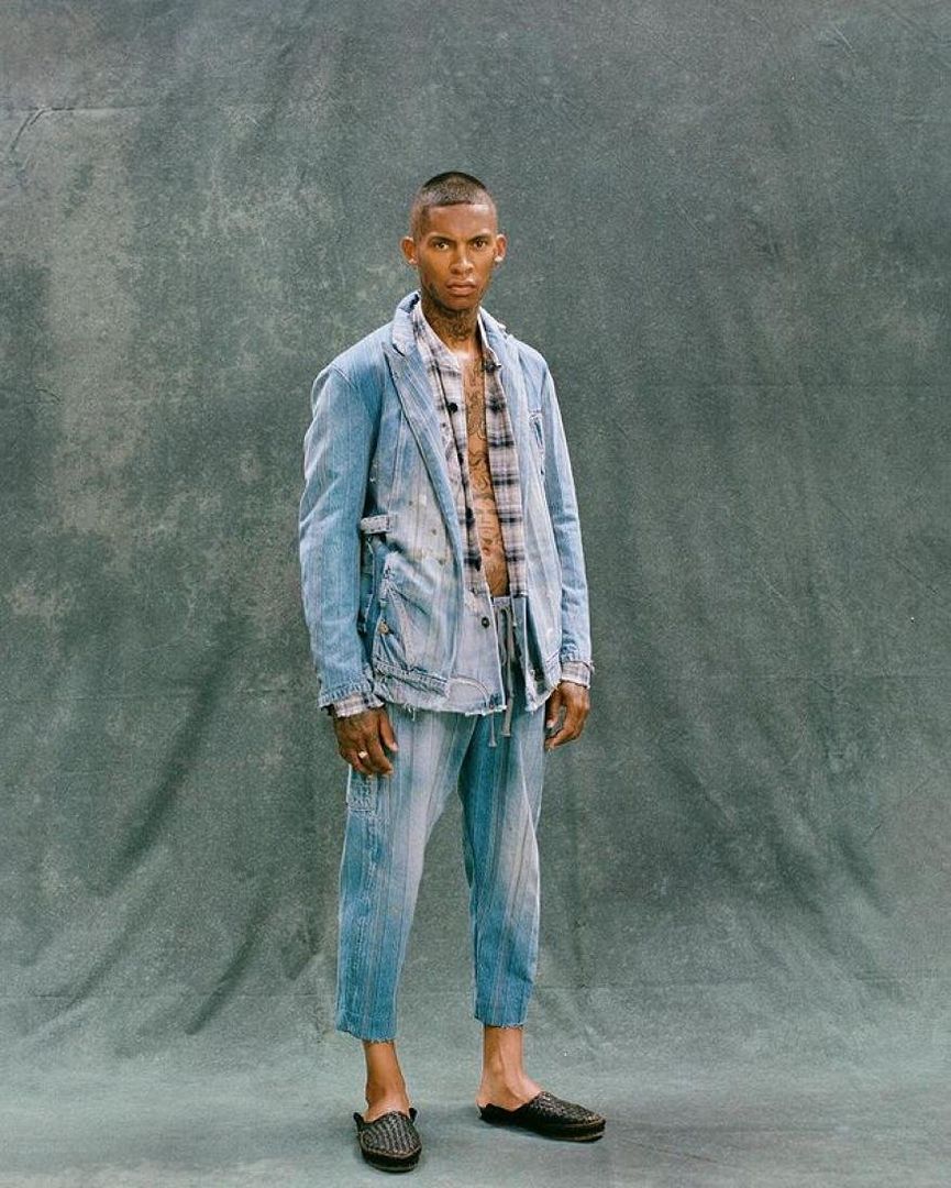 Portrait of a man in a light-blue denim suit, with a plaid shirt, posing against a gray backdrop in a fashion or editorial setting.