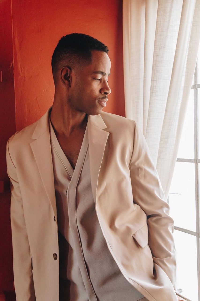 Portrait of a Black man in a tan suit standing by a window with natural light. He is looking off to the side.