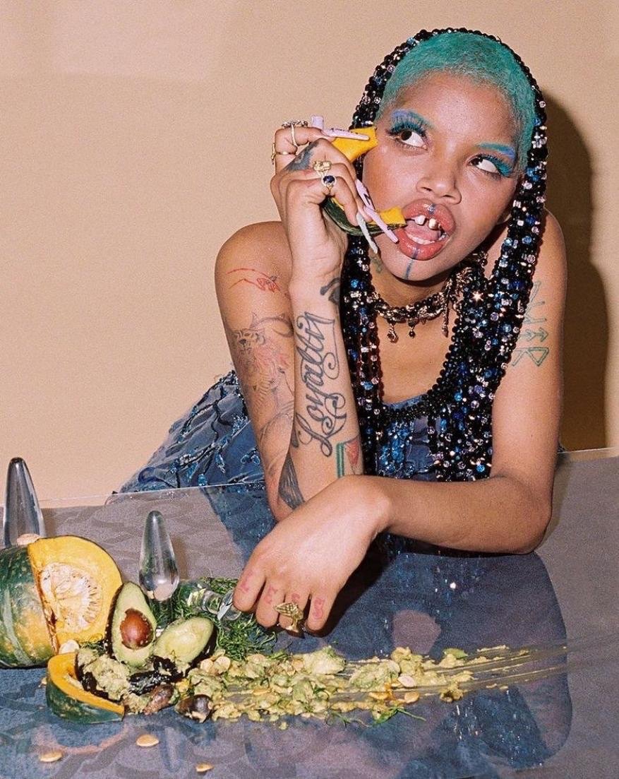 Woman poses at table with chopped squash and avocados, teal braided hair, creative makeup, and bejeweled top against tan background.