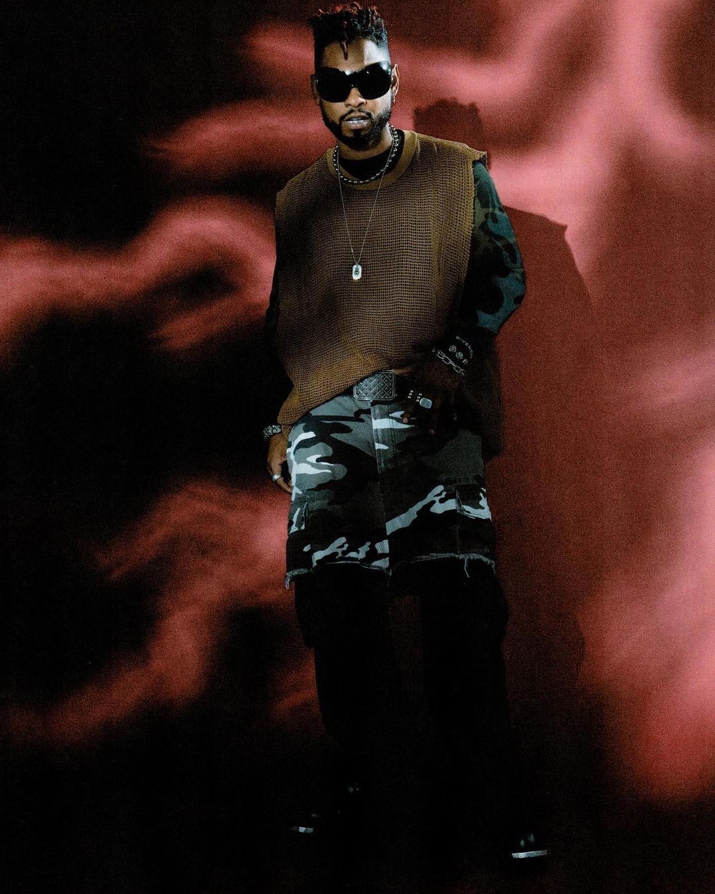 Adult male wearing sunglasses, vest, and camouflage pants poses against a dark backdrop with red smoke effect, looking forward.