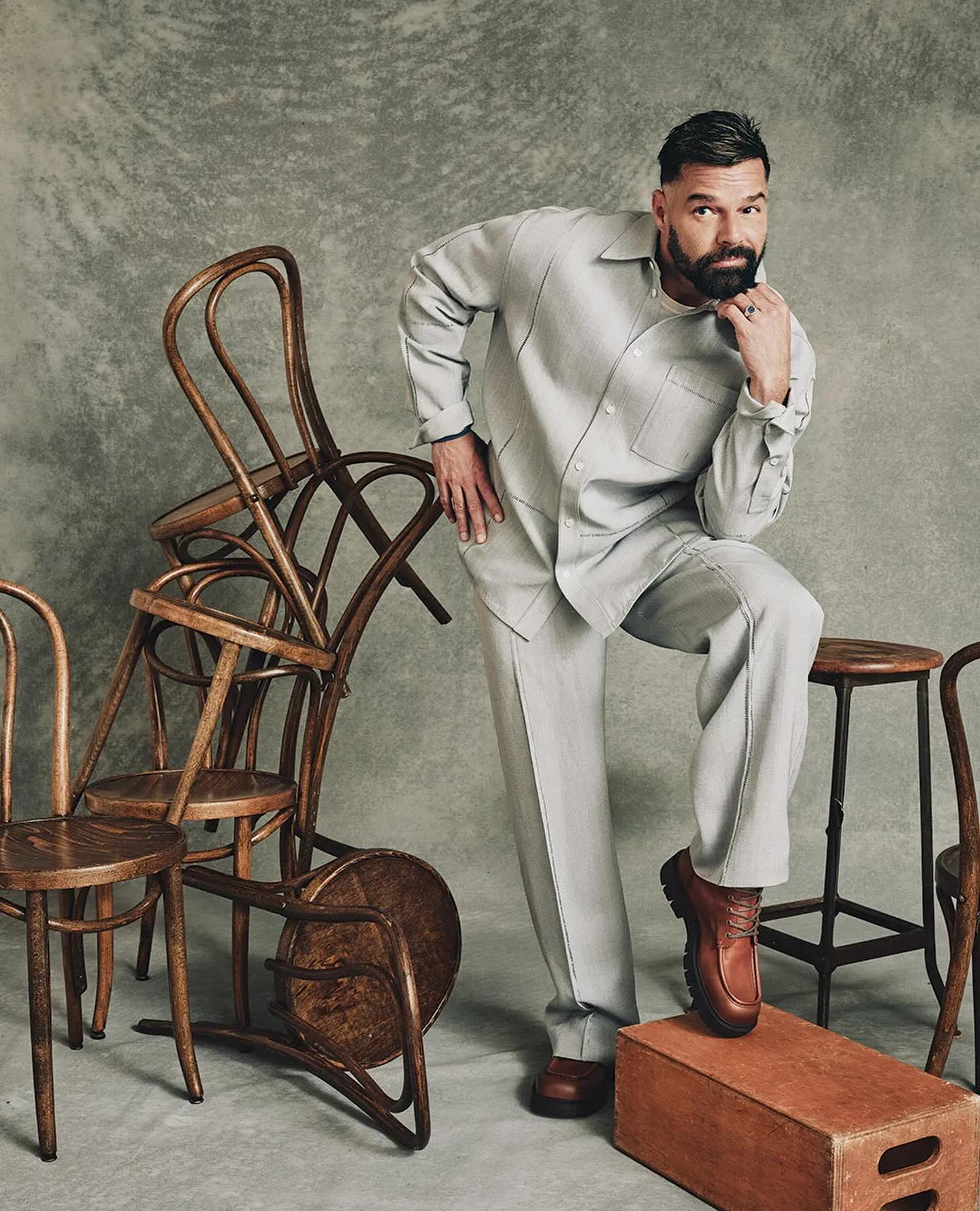 Bearded man in a grey suit poses indoors surrounded by wooden chairs and a stool, looking at the camera with a serious expression.