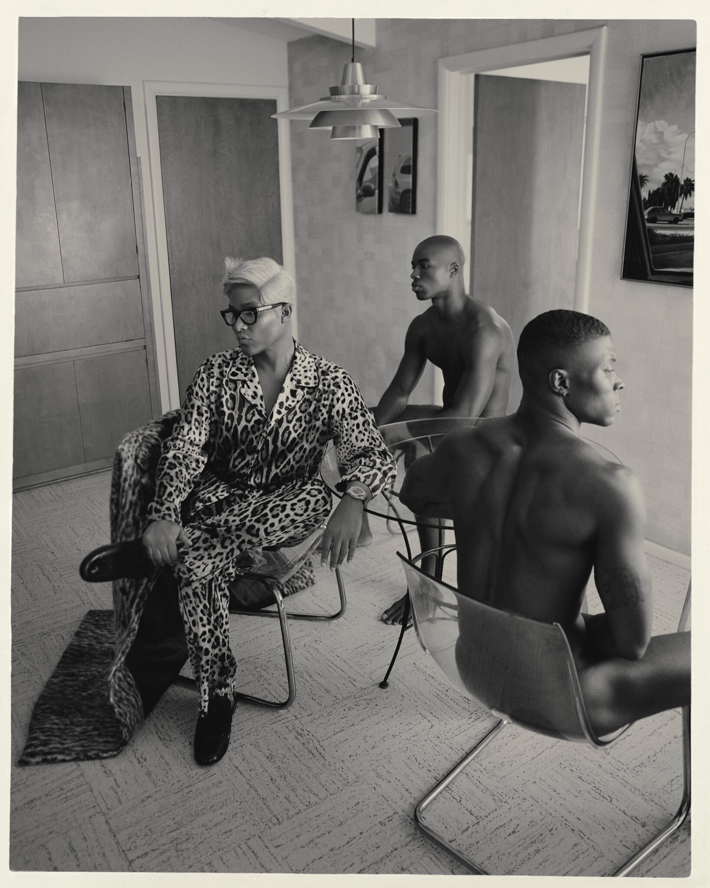 Three men are indoors. One is seated wearing a leopard-print pajama set and glasses, while two bare-chested men are seated behind him.