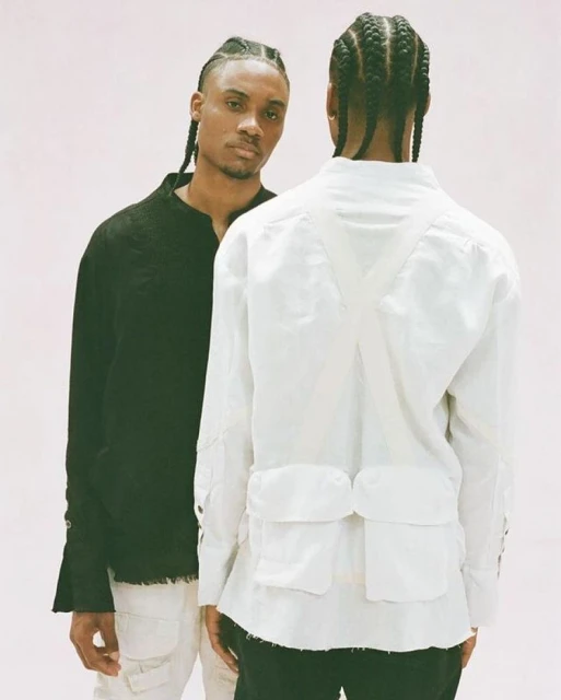 Two adult men with braided hair pose in a studio with a white backdrop. One man wears a black shirt, and the other wears a white utility jacket.