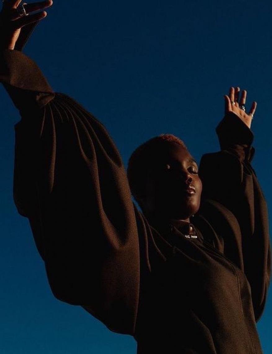 Low angle view of a Black woman with short pink hair posing with arms raised in front of a bright blue sky, wearing a brown long-sleeve shirt.