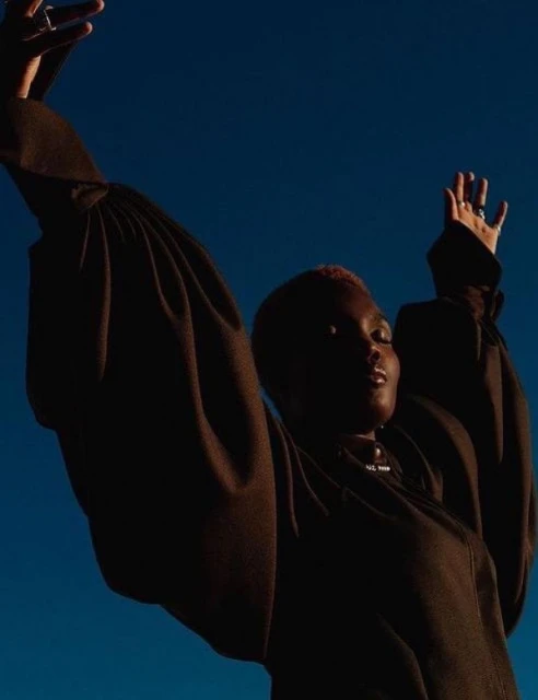 Low angle view of a Black woman with short pink hair posing with arms raised in front of a bright blue sky, wearing a brown long-sleeve shirt.