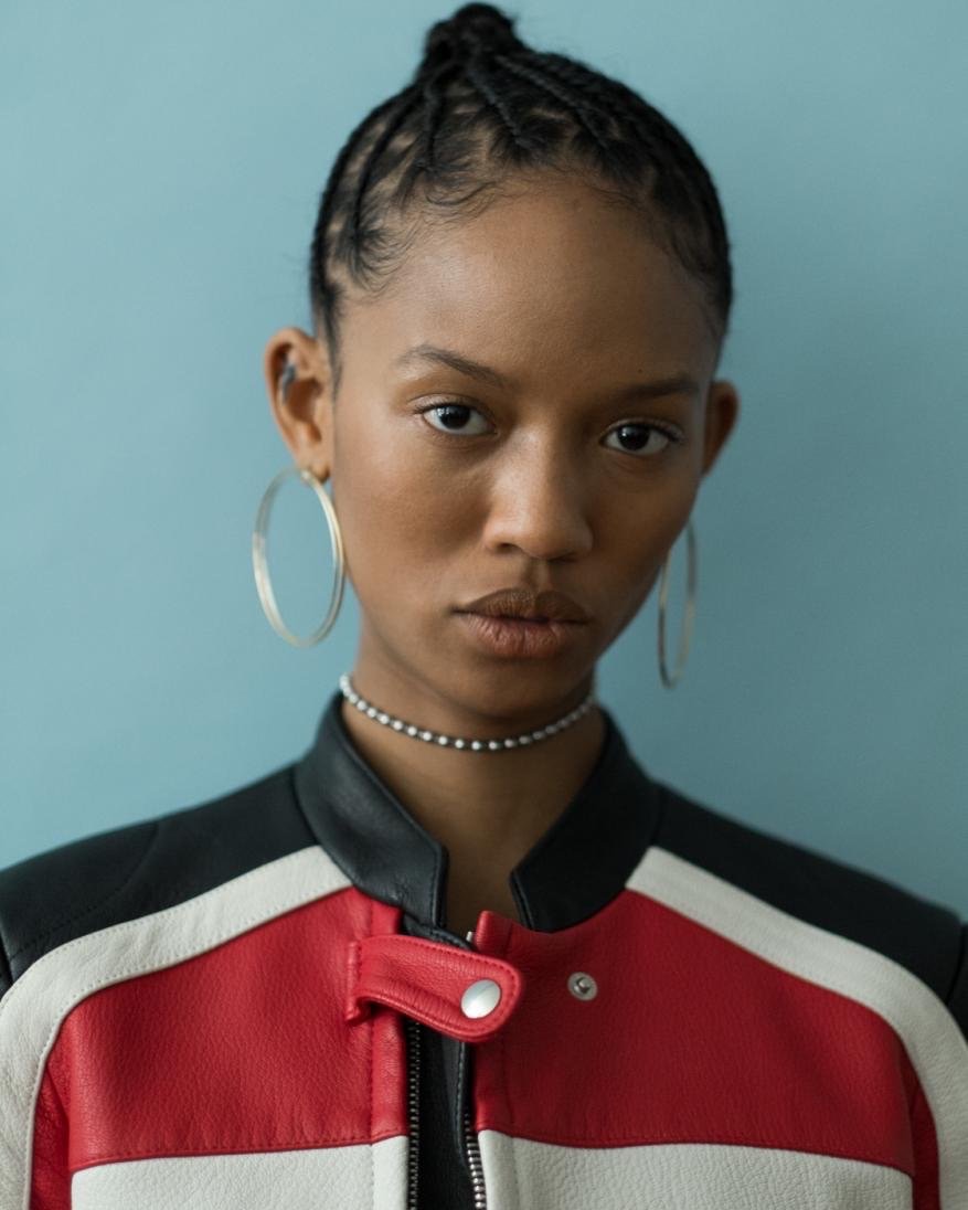 Close-up portrait of a stylish young woman with braided hair, silver hoop earrings, and a red, white, and black jacket against a blue background.