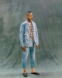 Man wearing a distressed light blue denim suit and plaid shirt standing against a textured grey backdrop.