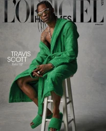 Travis Scott seated on a white stool wearing a textured green robe and matching green slippers against a grey seamless background