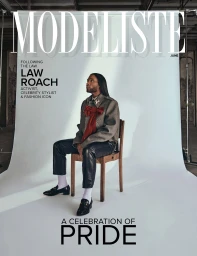 Law Roach seated on a wooden chair against a white seamless backdrop wearing a grey jacket and leather trousers