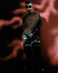 Man wearing sunglasses and a brown knit vest standing against a textured red and black patterned background