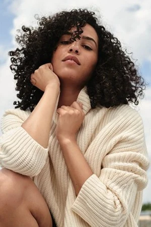 Portrait of a woman with dark curly hair wearing a cream turtleneck sweater against a clear sky background