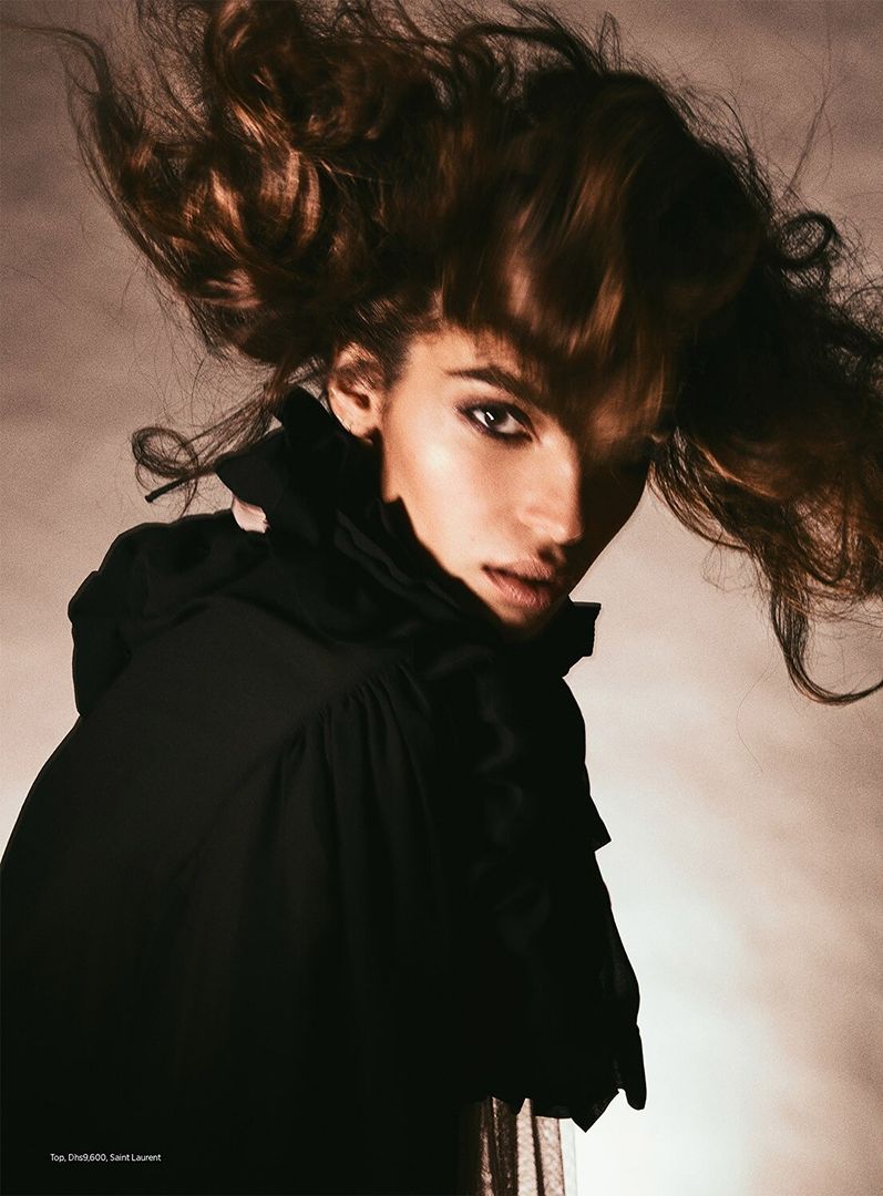 Portrait of a woman with dramatic hair and makeup wearing a black top with ruffled collar in a studio environment with soft lighting.