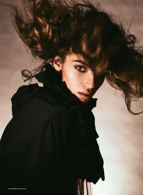 Portrait of a woman with dramatic hair and makeup wearing a black top with ruffled collar in a studio environment with soft lighting.