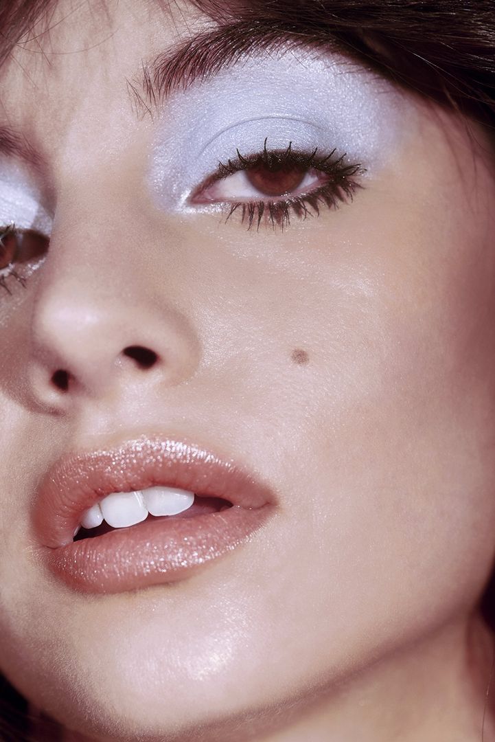 Close-up portrait of a woman featuring pale blue eyeshadow, dark mascara on lashes, and glossy lips with a noticeable mole on her cheek.