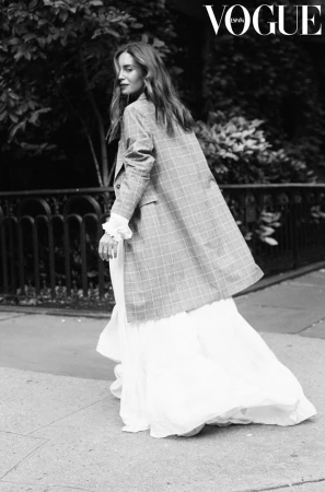 Black and white editorial fashion shot of a woman in a plaid blazer and long white ruffled dress walking outdoors.