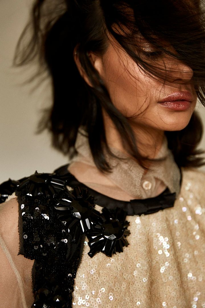 Close-up of an adult female model with dark hair, wearing a sequined top with beaded details, against an off-white backdrop in a studio shot.