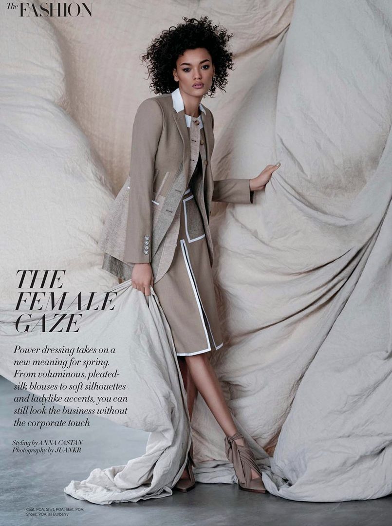Fashion editorial image featuring an adult female model in a tailored beige skirt suit, posing against a draped beige background.