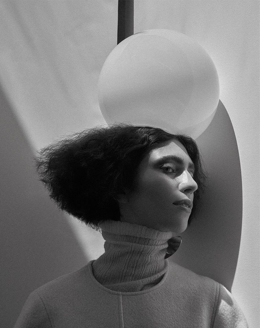 Monochromatic portrait of a woman with curly hair wearing a turtleneck, lit with natural light, featuring an overhead light fixture.